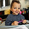 child, toddler, smiling, crayon, drawing, art, table, striped_shirt, indoor, living_room, cozy, face, happy, playful, person, scribbles, paper, toy, background, young