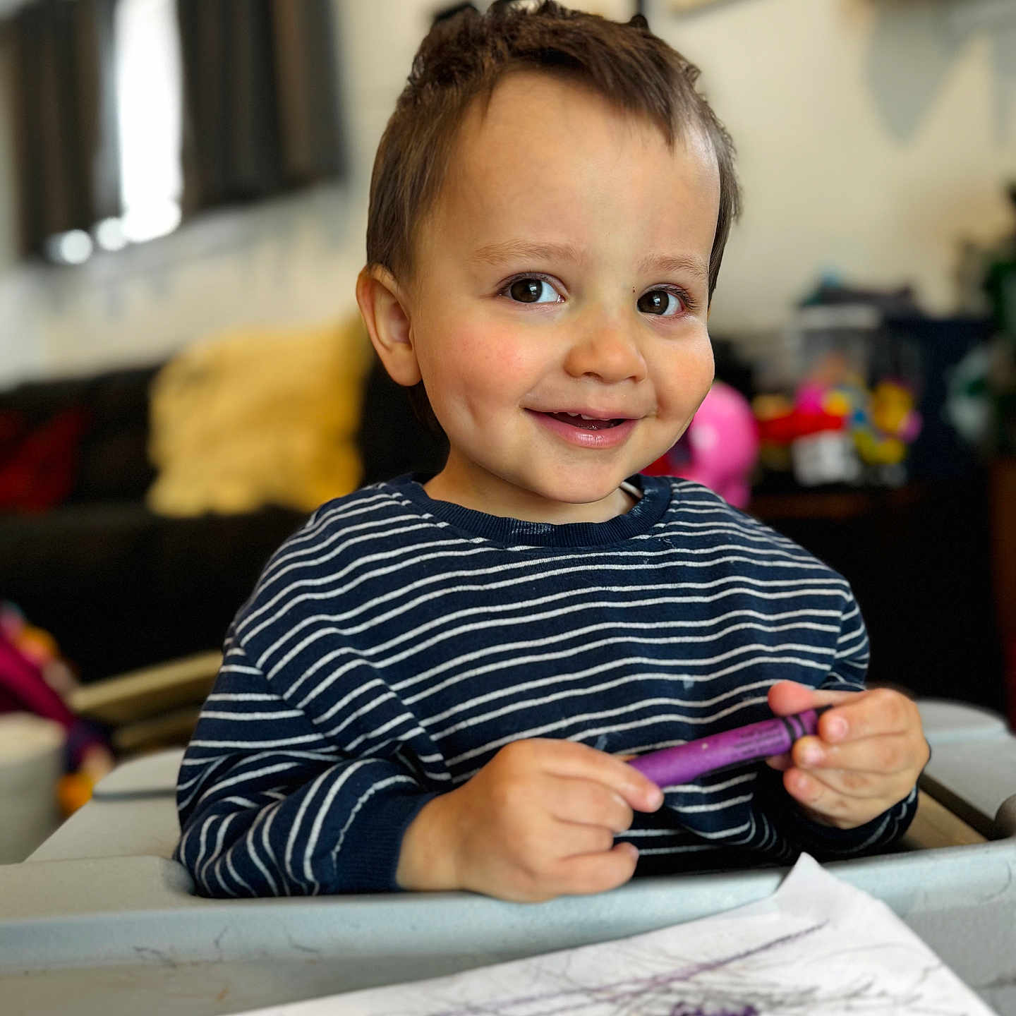 Eli is registered to the contest to win money with this photo: child, toddler, smiling, crayon, drawing, art, table, striped_shirt, indoor, living_room, cozy, face, happy, playful, person, scribbles, paper, toy, background, young