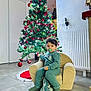 armchair, child, christmas_tree, cozy, curly_hair, festive, floor_tiles, green_sweater, holiday, holiday_decorations, home, indoor, lights, pacifier, radiator, red_ornaments, smiling, socks, toddler, young_child