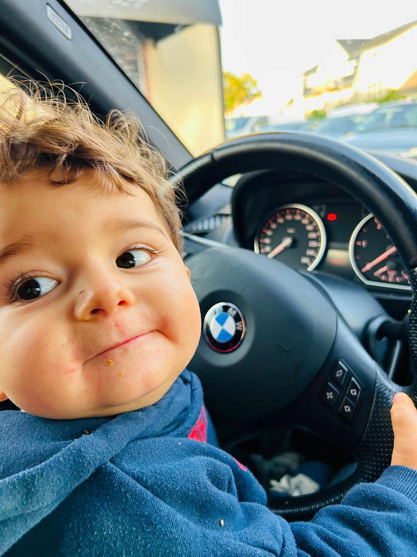 Léandro participe au concours pour gagner de l'argent avec cette photo : child, toddler, face, curly_hair, steering_wheel, car_interior, bmw_logo, blue_hoodie, hand, dashboard, speedometer, daylight, window, parked_cars, street, expression, portrait, messy_face, playful, indoors