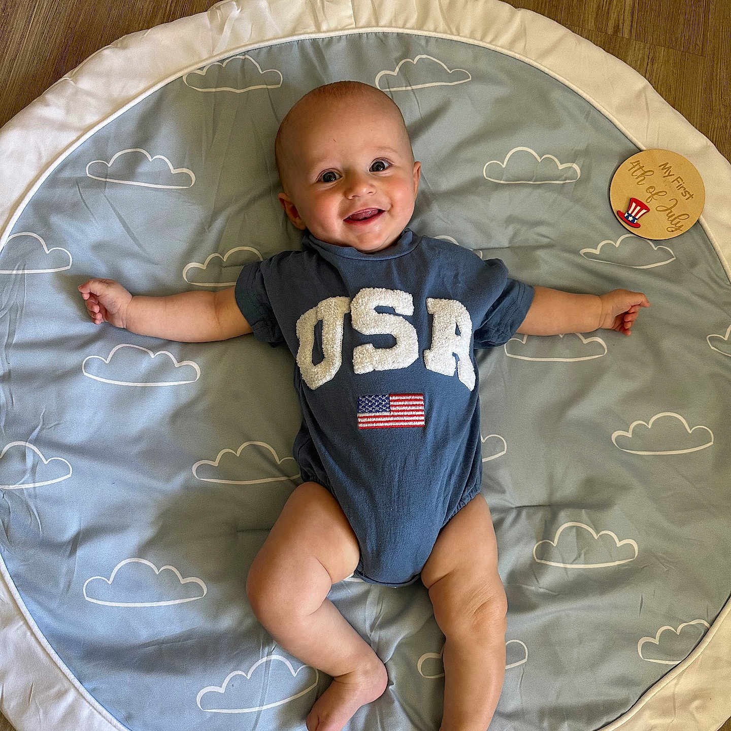 Grayson joined the competition — help win amazing prizes! american_flag, arms_outstretched, baby, blue_clothing, child, cloud_pattern, cute, floor, happy, head, indoor, infant, legs_bent, lying_down, onesie, playmat, portrait, smiling, usa, wooden_floor