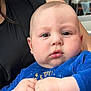 baby, child, infant, blue_clothing, hands, face, chubby_cheeks, indoor, person, adult, black_clothing, portrait, closeup, curious_expression, skin, human, holding, seated, candid, soft_light