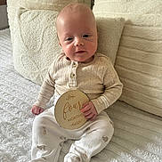 Marley is registered to the contest to win money with this photo: baby, infant, child, four_months, milestone, sign, wooden_sign, sitting, blanket, pillows, bed, clothing, onesie, footed_pajamas, face, hand, indoors, soft_lighting, cute, portrait