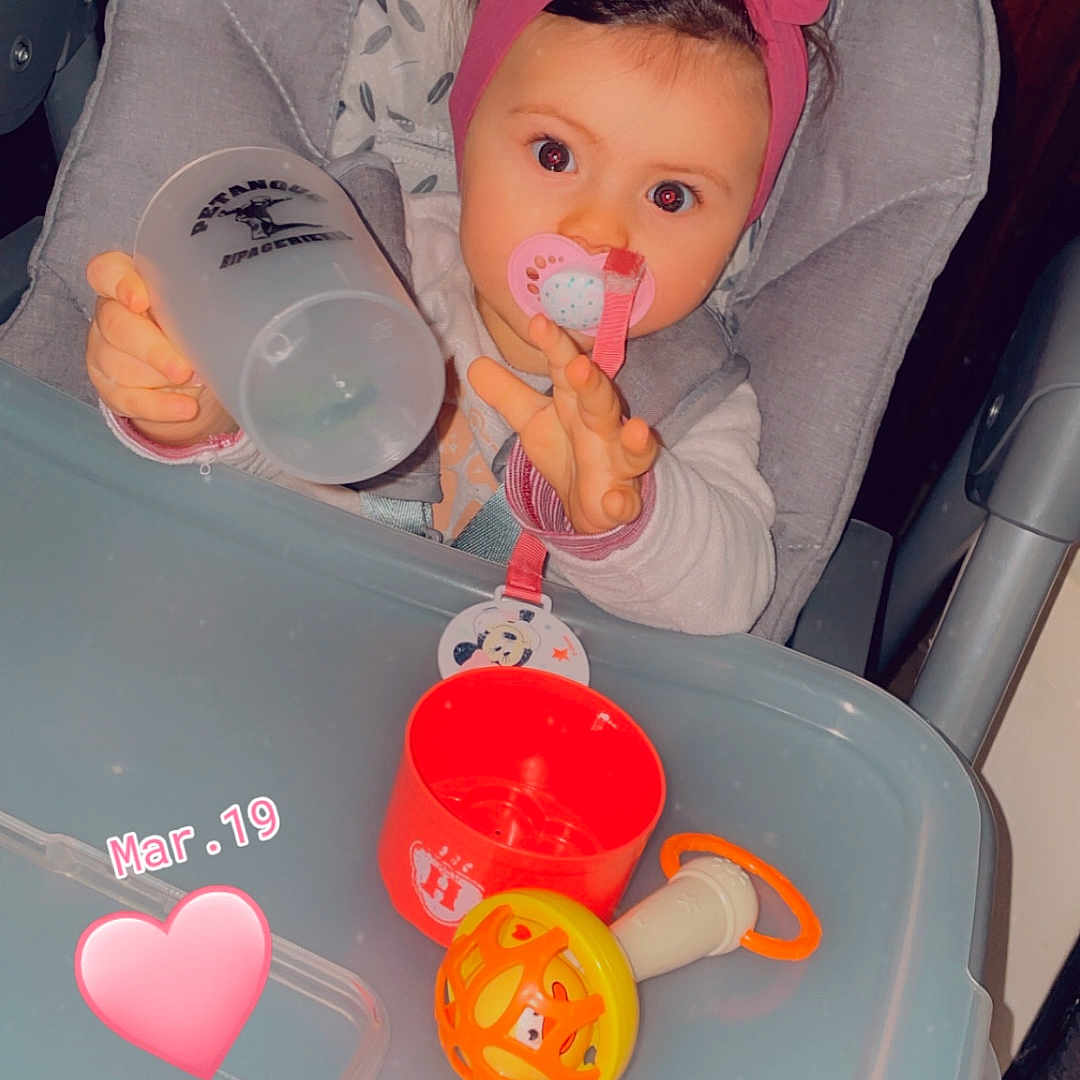 Lina participe au concours pour gagner de l'argent avec cette photo : baby, high_chair, bottle, headband, pink_bow, toy, pacifier, child, indoor, table, face, hand, seat, cute, infant, expression, light, plastic, red, gray