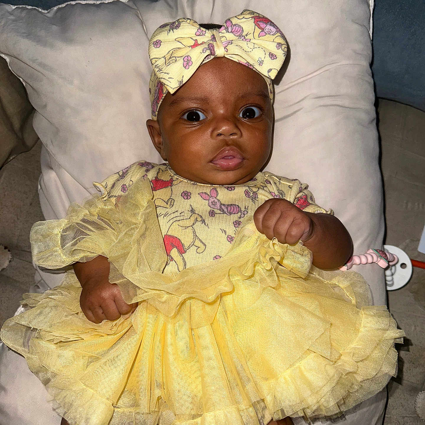 Olympia is registered to the contest to win money with this photo: baby, child, clothing, cushion, cute, dress, expression, fabric, face, hand, headband, indoors, infant, looking, person, pillow, portrait, sitting, soft, yellow_dress