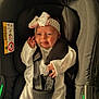 baby, infant, car_seat, headband, bow, white_clothing, straps, safety, cute, smiling, tongue_out, child, newborn, seat, black, green, fabric, comfort, indoors, portrait