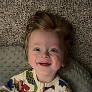 Dax is registered to the contest to win money with this photo: child, toddler, smiling, face, hair, onesie, dinosaur_pattern, baby, joyful, happy, indoor, carpet, pillow, portrait, cute, infant, clothing, expression, lying_down, human