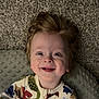 child, toddler, smiling, face, hair, onesie, dinosaur_pattern, baby, joyful, happy, indoor, carpet, pillow, portrait, cute, infant, clothing, expression, lying_down, human