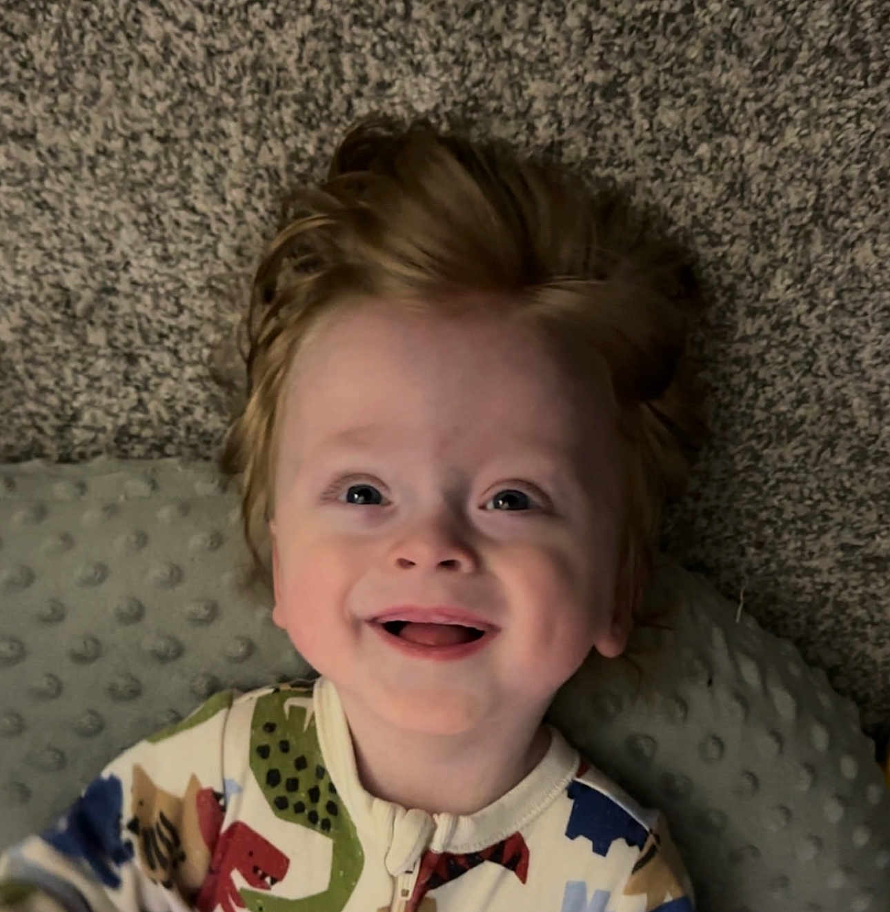 Dax is registered to the contest to win money with this photo: child, toddler, smiling, face, hair, onesie, dinosaur_pattern, baby, joyful, happy, indoor, carpet, pillow, portrait, cute, infant, clothing, expression, lying_down, human