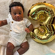 Khi is registered to the contest to win money with this photo: baby, child, balloon, number_three, white_overalls, socks, curly_hair, blanket, celebration, indoor, portrait, cute, infant, smiling, skin, person, happy, cozy, soft_texture, lighting