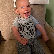Gabriel is registered to the contest to win money with this photo: baby, child, smiling, tongue_out, gray_shirt, text_on_clothing, pants, armchair, beige_furniture, happy, indoor, person, cute, playful, young_child, sitting, candid, portrait, home, funny_text