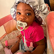 Daesha joined the competition — help win amazing prizes! baby, pacifier, bonnet, pink_shirt, socks, toy, playpen, baby_bottle, cushion, indoor, child, infant, cute, seated, person, closeup, face, expression, soft_lighting, cozy