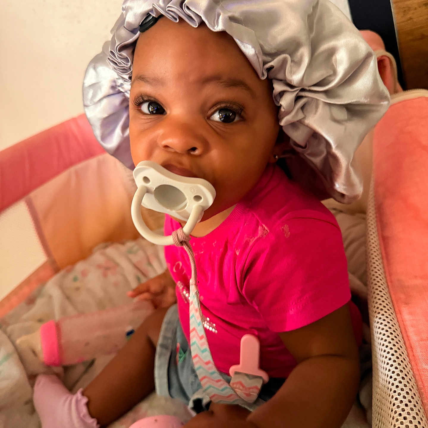 Daesha joined the competition — help win amazing prizes! baby, pacifier, bonnet, pink_shirt, socks, toy, playpen, baby_bottle, cushion, indoor, child, infant, cute, seated, person, closeup, face, expression, soft_lighting, cozy