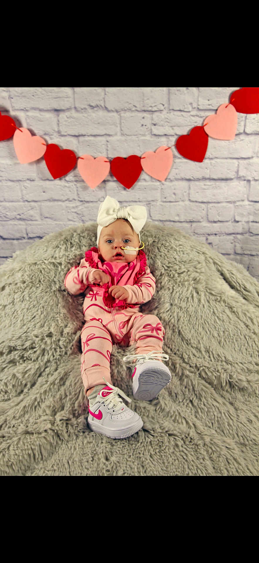 Indy is registered to the contest to win money with this photo: baby, infant, child, headband_bow, feeding_tube, onesie, scarf, sneakers, plush_blanket, heart_garland, brick_wall, portrait, studio_shot, sitting, pink_outfit, white_bow, cute, tiny_shoes, soft_texture, decor