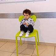 Zayn participe au concours pour gagner de l'argent avec cette photo : bulletin_board, casual_clothing, child, curious_expression, curly_hair, floor_tile, furniture, green_chair, indoor, paper, person, room, shoe, shoes, sitting, tile_floor, waiting_area, wall, white_shoes, young_child