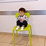 bulletin_board, casual_clothing, child, curious_expression, curly_hair, floor_tile, furniture, green_chair, indoor, paper, person, room, shoe, shoes, sitting, tile_floor, waiting_area, wall, white_shoes, young_child