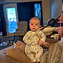 baby, infant, person, onesie, indoor, living_room, couch, seated, curious, face, hand, adult, tattoo, furniture, window, curtains, soft_lighting, home, cozy, casual
