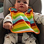 baby, infant, bib, rainbow_bib, onesie, highchair, seat, safety_strap, chubby_cheeks, clenched_fist, neutral_expression, curtain, wood_panel, indoors, portrait, closeup, pajamas, bear_print, colorful, adorable