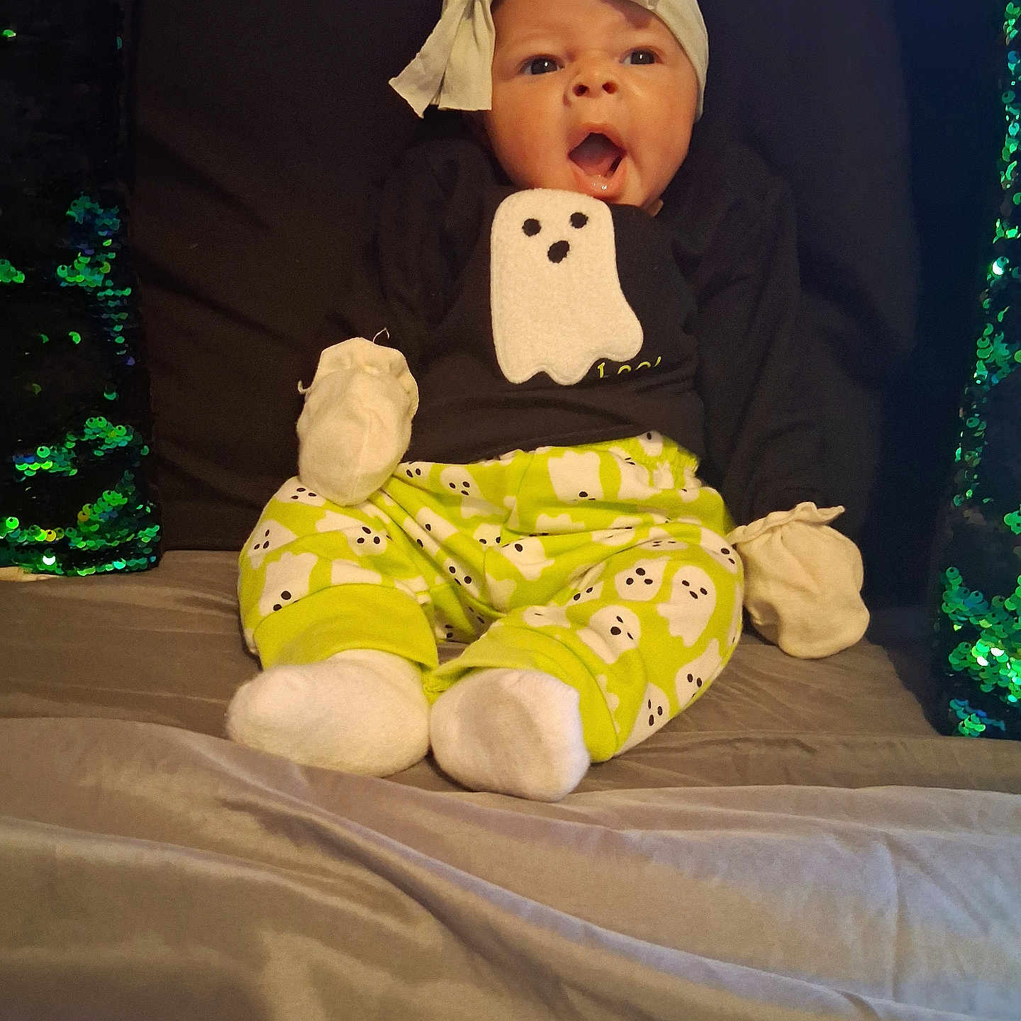 Ma'Kenna is registered to the contest to win money with this photo: baby, bed, black_shirt, child, clothing, comfortable, cute, ghost_pattern, green, headwrap, indoors, infant, mittens, open_mouth, pillow, portrait, sequin_pillow, sitting, socks, surprised_expression