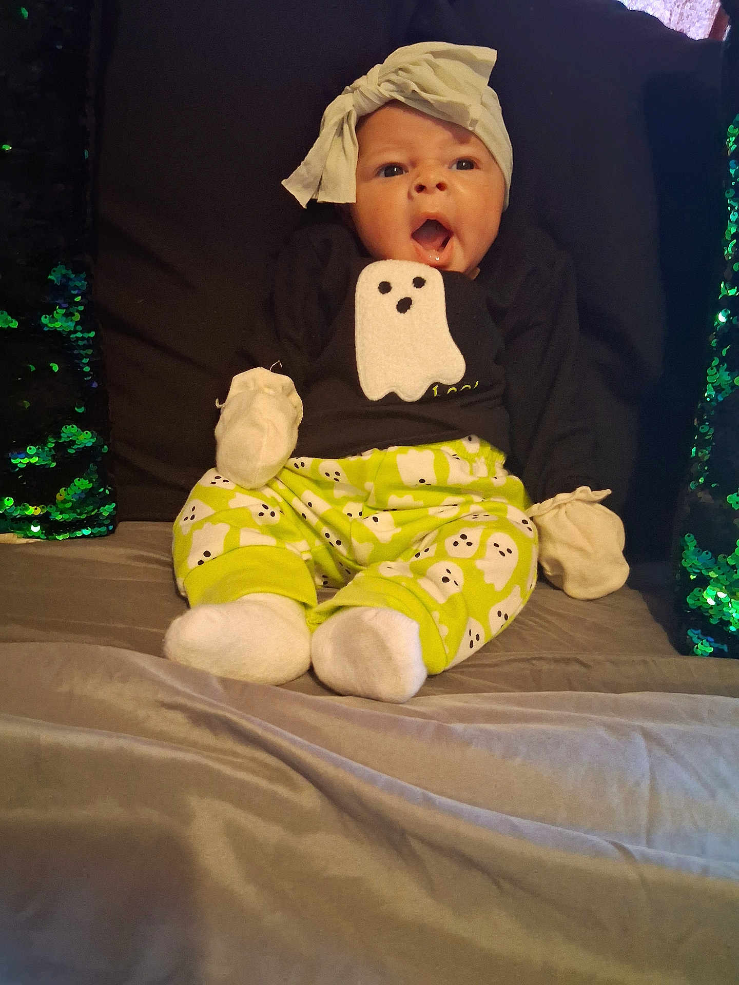 Ma'Kenna is registered to the contest to win money with this photo: baby, child, infant, ghost_pattern, headwrap, mittens, socks, green, black_shirt, bed, pillow, sequin_pillow, surprised_expression, open_mouth, sitting, indoors, cute, comfortable, clothing, portrait