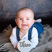 Tav is registered to the contest to win money with this photo: baby, blanket, blue_eyes, child, cute, denim_overalls, face, happy, head, indoor, infant, laying_down, person, portrait, skin, smiling, three_months, white_shirt, wooden_sign, young