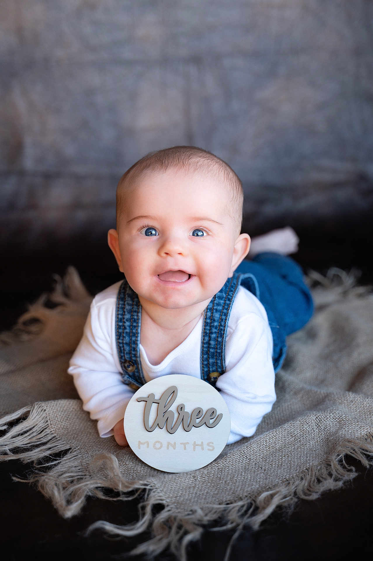 Tav is registered to the contest to win money with this photo: baby, blanket, blue_eyes, child, cute, denim_overalls, face, happy, head, indoor, infant, laying_down, person, portrait, skin, smiling, three_months, white_shirt, wooden_sign, young