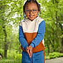 child, glasses, braids, beads, smile, hoodie, pants, outdoor, forest, trees, grass, pathway, nature, happy, person, portrait, casual_clothing, standing, daylight, greenery