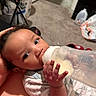 baby, bottle, feeding, infant, hand, onesie, indoor, carpet, laundry_basket, gallon_jug, floor, person, closeup, cute, child, expression, holding, skin, face, young
