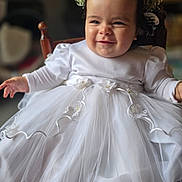 Alya participe au concours pour gagner de l'argent avec cette photo : baby, child, white_dress, flower_crown, smile, cute, sitting, chair, indoor, portrait, celebration, formal_wear, footwear, happy, young_child, tulle, cute_expression, headwear, person, infant