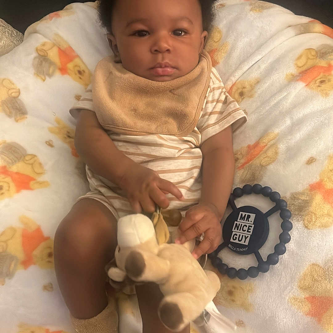 Messiah is registered to the contest to win money with this photo: baby, bib, blanket, calm, child, comfort, cozy, cute, indoor, infant, nursery, person, plush_toy, seated, socks, soft_texture, striped_clothing, teddy_bear_print, toy, young_child