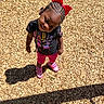 black_tshirt, braided_hair, casual_clothing, child, cute, daylight, fun, girl, happy, outdoor, person, pink_shoes, pink_shorts, playground, red_bow, shadow, smile, standing, sunny, wood_chips