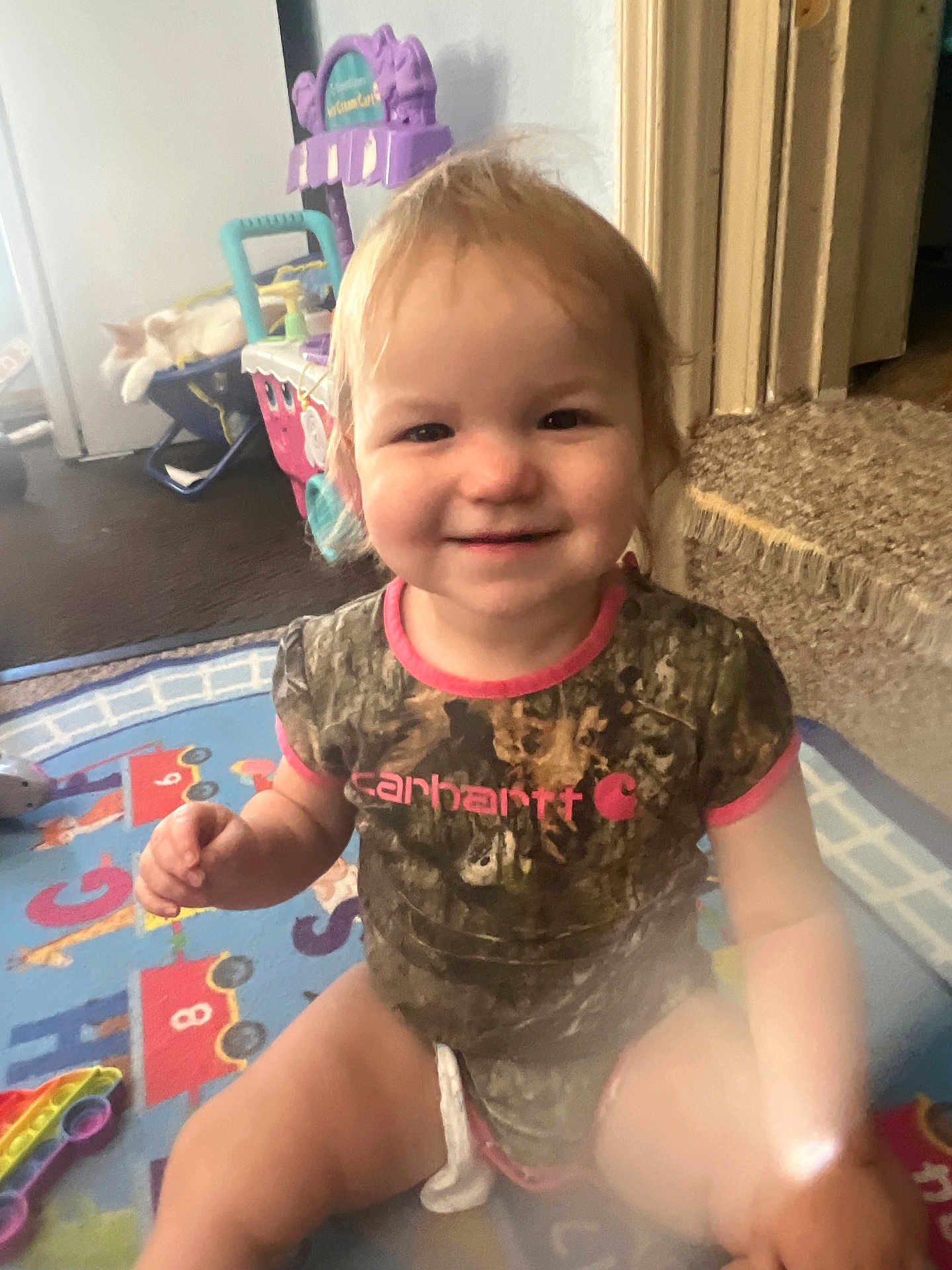 Nova is registered to the contest to win money with this photo: toddler, child, smiling, indoor, play_mat, toy, cat, couch, carhartt, camouflage, person, happy, sitting, furniture, floor, carpet, light, hair, cute, baby