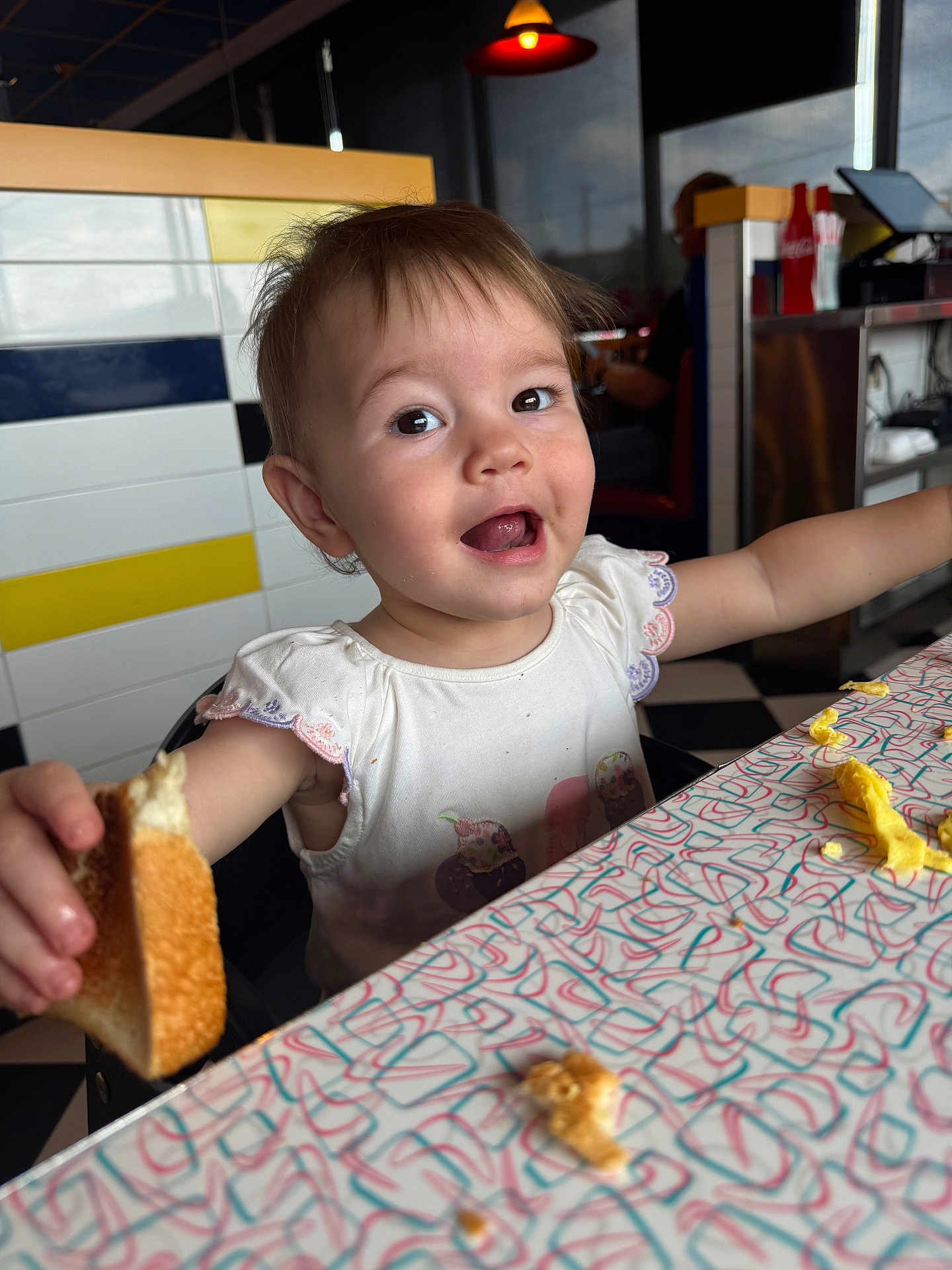 Emily joined the competition — help win amazing prizes! toddler, child, bread, food, table, smile, happy, mealtime, diner, scrambled_eggs, crumbs, white_shirt, indoor, person, face, cute, expression, colorful, highchair, eating