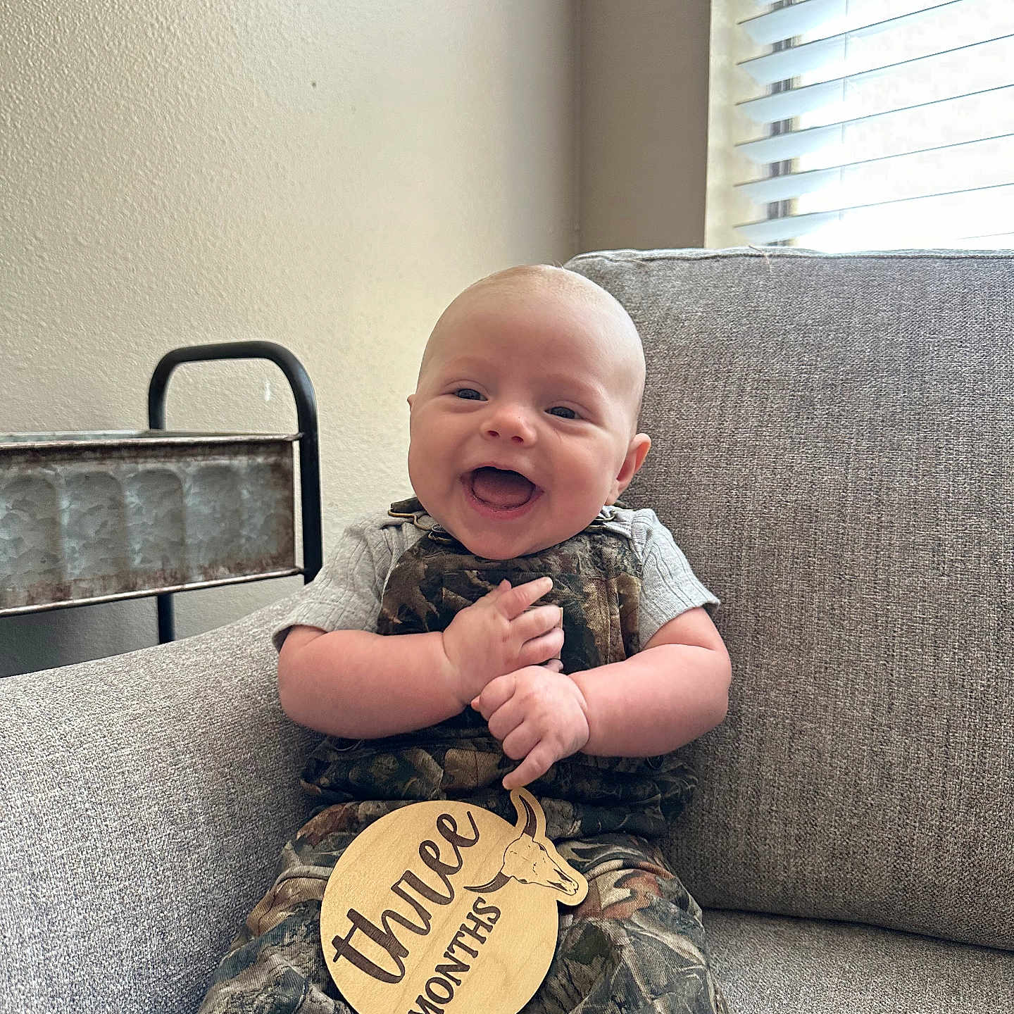 Kacey joined the competition — help win amazing prizes! baby, blinds, camo_overalls, child, clothing, couch, face, hands, happy, indoor, infant, light, person, portrait, sign, sitting, smiling, three_months, window, wooden_sign