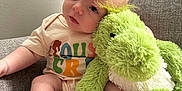 Kacey is registered to the contest to win money with this photo: baby, plush_toy, dinosaur, couch, infant, foot, shirt, sitting, indoor, cute, child, soft_toy, relaxed, head_tilt, colorful_text, furniture, light, window, wall, cozy