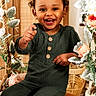 child, toddler, smiling, green_outfit, indoor, flowers, wooden_bench, happy, cute, portrait, curly_hair, buttoned_shirt, playful, decor, plant_leaves, bright, warm_lighting, face, person, sitting
