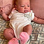 baby, infant, onesie, pink_socks, patterned_blanket, pillow, face, tongue_out, lying_down, cute, child, soft_lighting, indoors, newborn, human, skin, expression, clothing, bed, resting