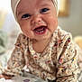 baby, smiling, face, headband, bow, eyes, sweater, patterned_clothing, infant, cute, happy, portrait, child, indoors, blanket, hand, closeup, cheerful, soft_light, young