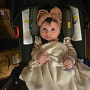 Elena is registered to the contest to win money with this photo: baby, child, stroller, blanket, bow, indoor, infant, clothing, person, car_seat, headwear, looking_up, curious, black_stroller, wood_floor, box, shoe_rack, carpet, patterned_rug, dim_lighting