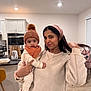 Neev is registered to the contest to win money with this photo: baby, woman, indoor, kitchen, warm_clothing, hat, pom_pom, sweater, headscarf, holding, smile, portrait, modern, furniture, countertop, appliance, person, child, cozy, happy