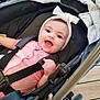 baby, infant, stroller, headband, bow, pink_clothing, smiling, happy, child, seatbelt, black_stroller, indoor, wooden_floor, cute, person, portrait, young_child, baby_girl, baby_clothes, baby_smile