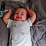baby, smiling, onesie, quilted_blanket, lying_down, happy, infant, child, face, arms_up, bedroom, soft_lighting, indoor, cute, person, sleepwear, patterned_clothing, head, toddler, relaxed