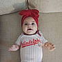 baby, infant, red_headband, bow, onesie, baseball, philadelphia, smiling, happy, couch, beige, textured, clothing, indoor, person, face, portrait, hand, cute, child