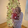 baby, woman, christmas_tree, holiday, festive, ornaments, lights, indoor, smiling, holding, joyful, celebration, family, red_clothing, headband, wood_floor, window_blinds, portrait, together, happy