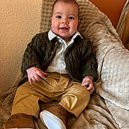 Jules a rejoint le concours — aidez-le/la à gagner de superbes lots ! baby, child, smile, clothing, pants, shoes, cardigan, blanket, couch, face, person, indoors, furniture, happy, cute, portrait, seated, warm_colors, cozy, young_child