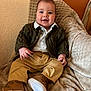 baby, child, smile, clothing, pants, shoes, cardigan, blanket, couch, face, person, indoors, furniture, happy, cute, portrait, seated, warm_colors, cozy, young_child