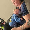 arm_tattoo, baby, blue_clothing, candid, casual, child, family, furniture, holding, home, indoor, peaceful, person, portrait, relaxed, resting, seated, sleeping, tattoo, woman