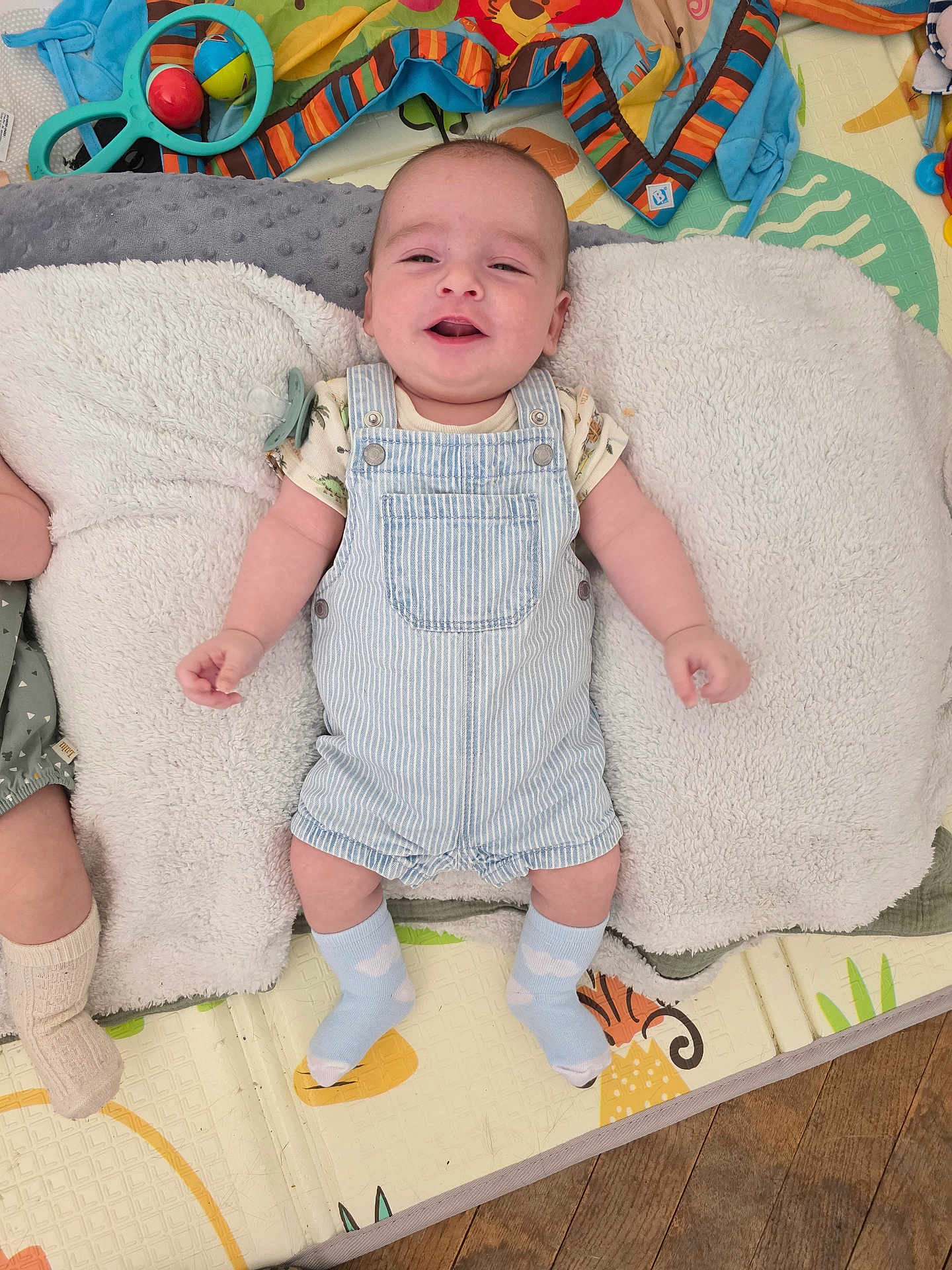 Charli a rejoint le concours — aidez-le/la à gagner de superbes lots ! baby, child, smiling, blue_overalls, socks, blanket, toys, play_mat, floor, happy, infant, indoor, cute, lying_down, patterned, striped_clothing, wooden_floor, paciifier, colorful, soft_texture