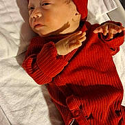 Ezra is registered to the contest to win money with this photo: baby, newborn, infant, red_clothing, hat, blanket, bed, sleepwear, human, skin, face, hand, foot, cute, indoors, lying_down, small, portrait, child, cozy