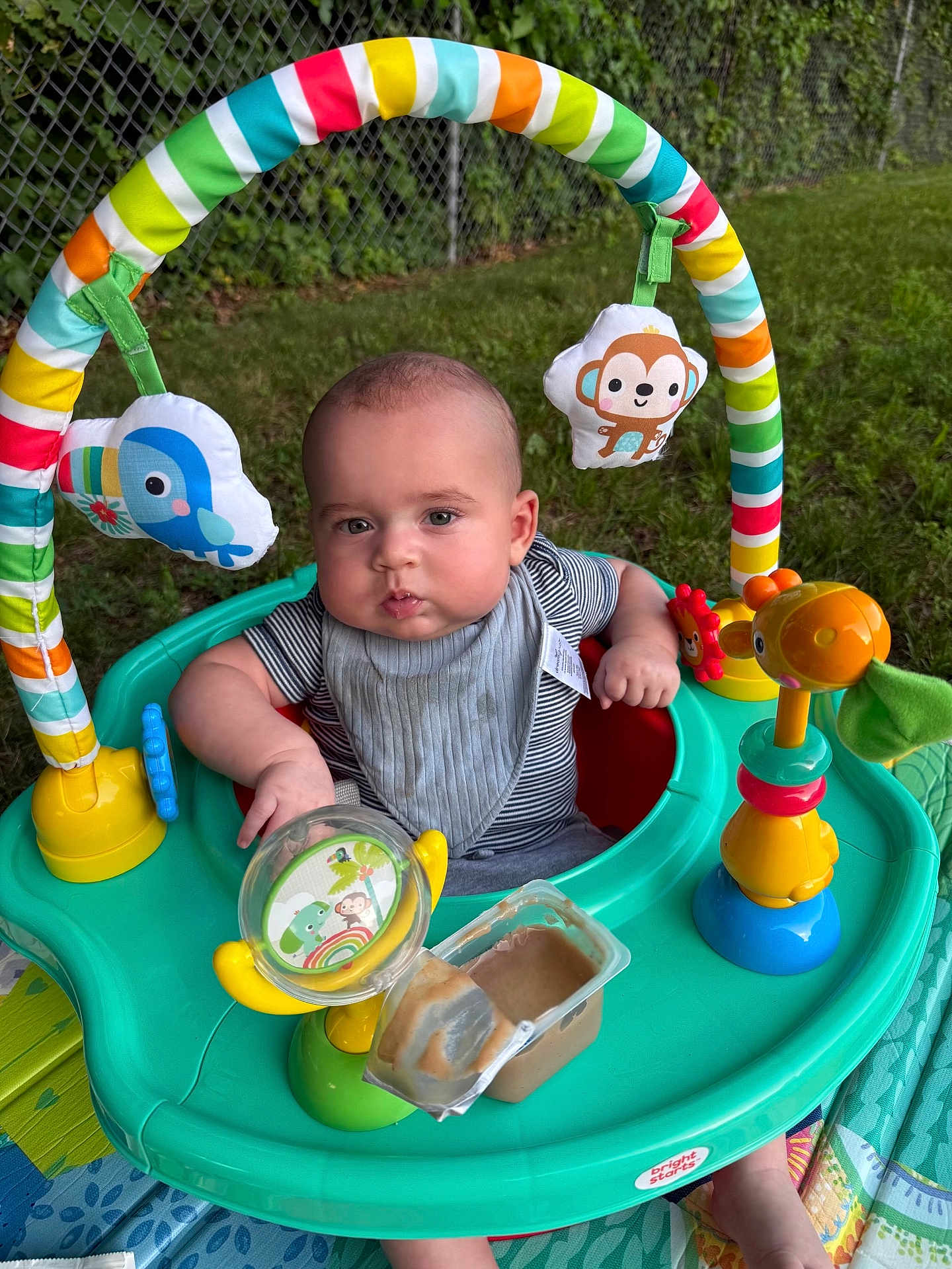 Dakeem is registered to the contest to win money with this photo: baby, infant, activity_center, toy, outdoor, grass, fence, colorful, play, child, feeding, bib, face, cute, person, seat, plastic, green, yellow, blue